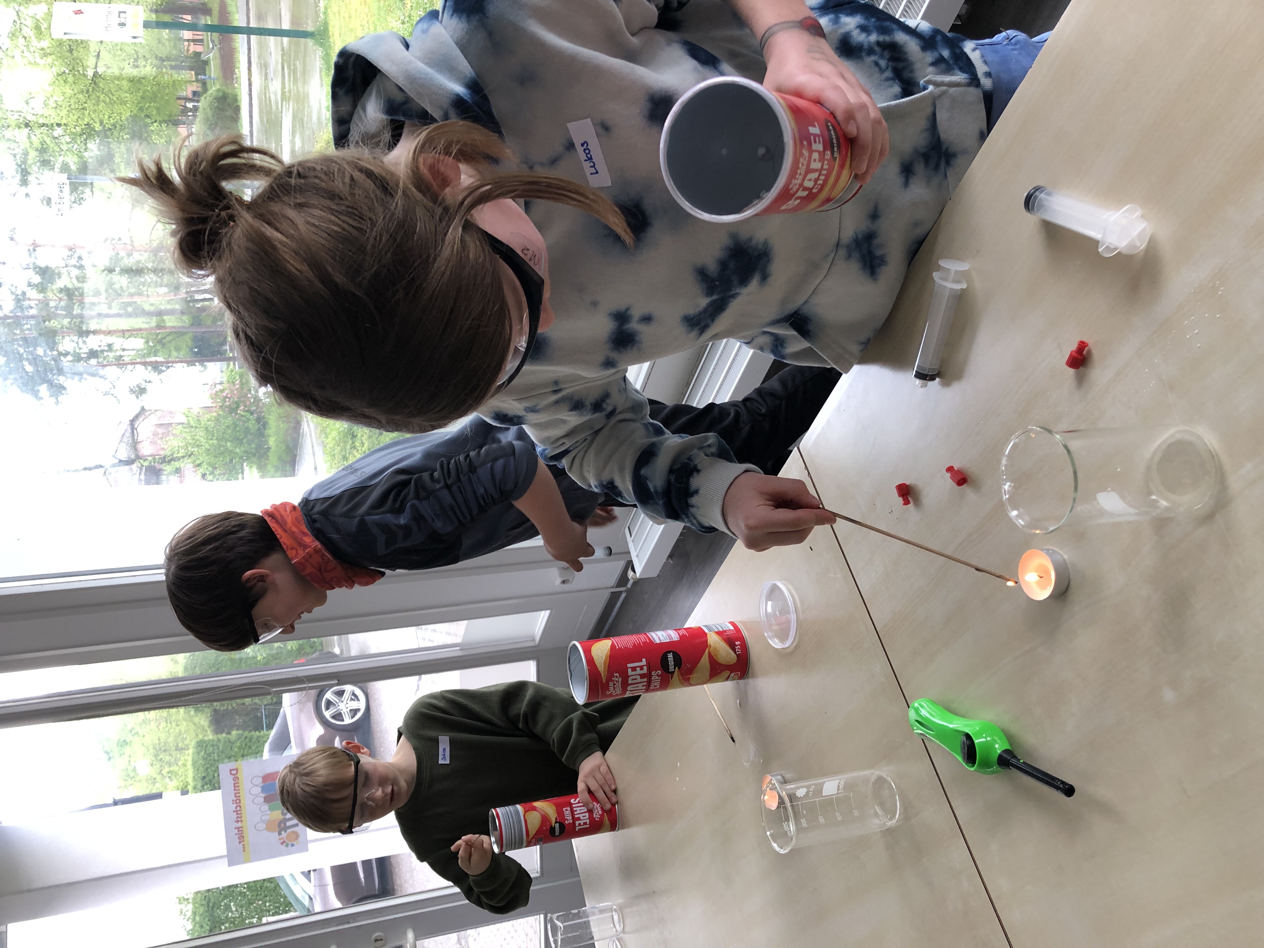  Drei Jungen mit Schutzbrillen experimentieren mit einer Kerze und Chipsdosen 