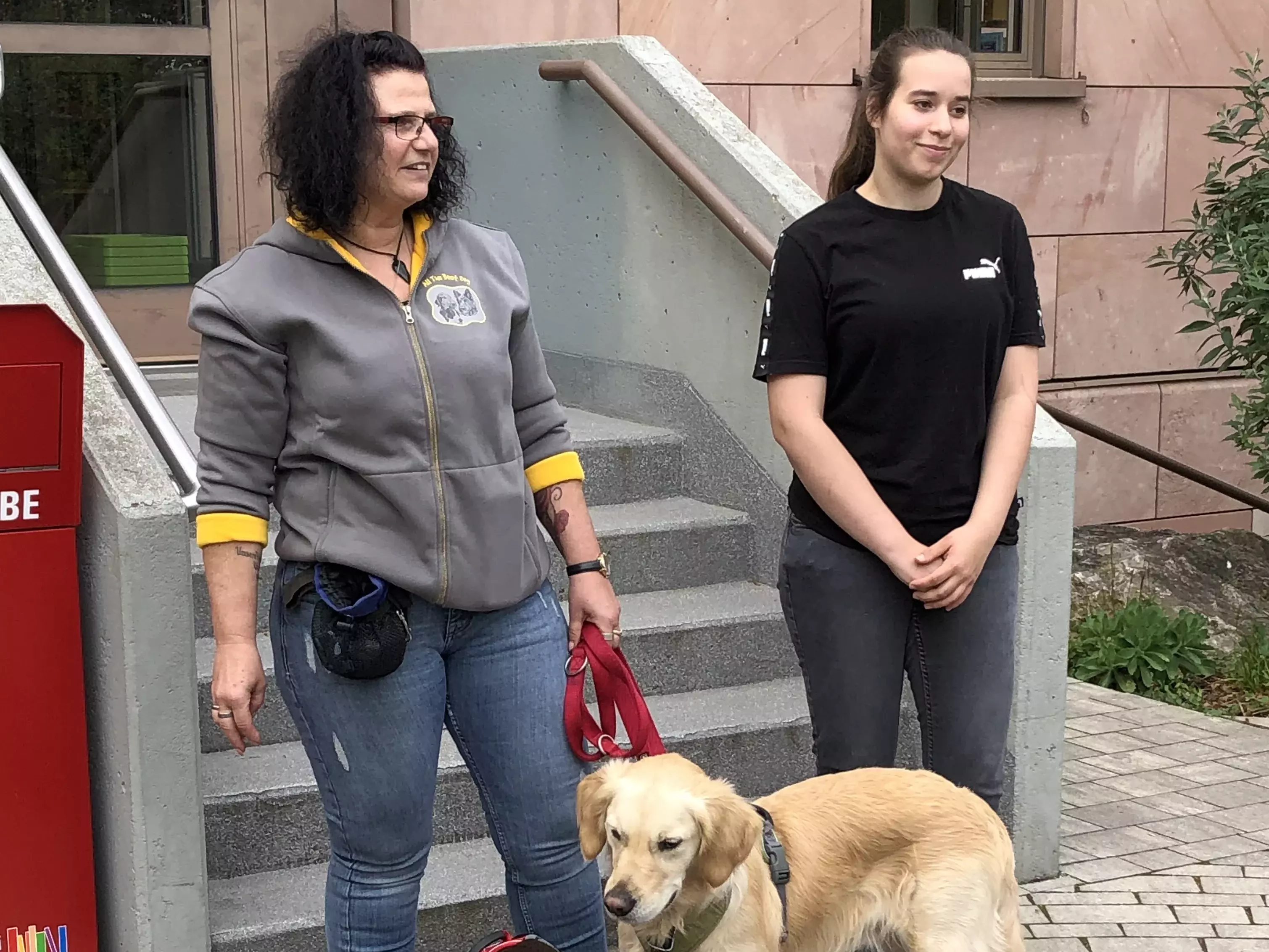 Carmen Nottrott und Auszubildene Leonie stehen it einem Hund vor dem Büchereieingang